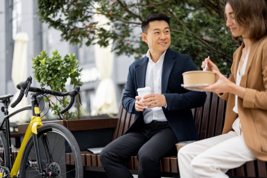 Asian businessman and caucasian businesswoman eating food and drinking coffee while having lunch at work. Concept of rest and break on job. Business people sitting on bench on city street
