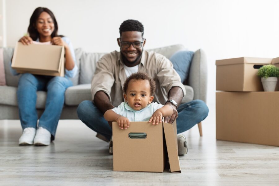 Moving, Housing and Real Estate Concept. Joyful black family of three people having fun, guy pushing child in carton box at new apartment. Happy African American parents celebrating relocation